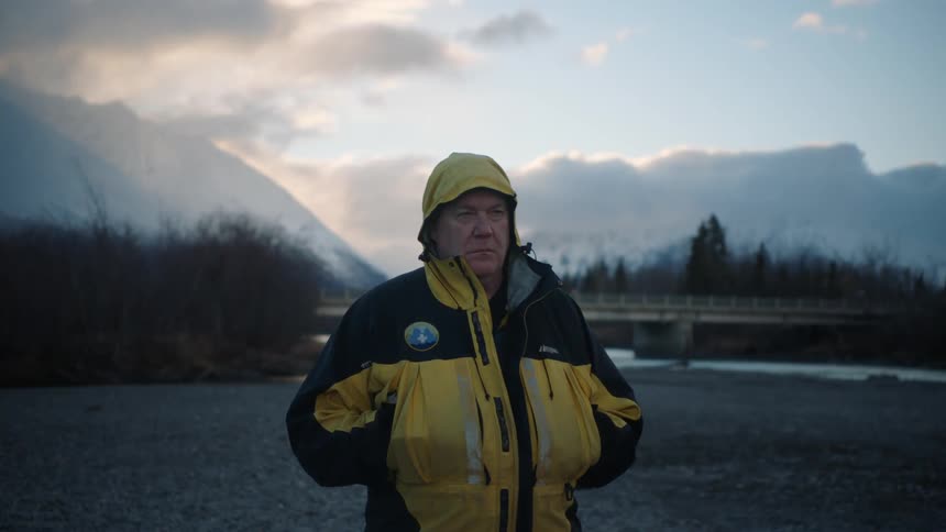 Man wearing a thick hooded yellow jacket stands in front of a bridge over a river in the mountains at morning