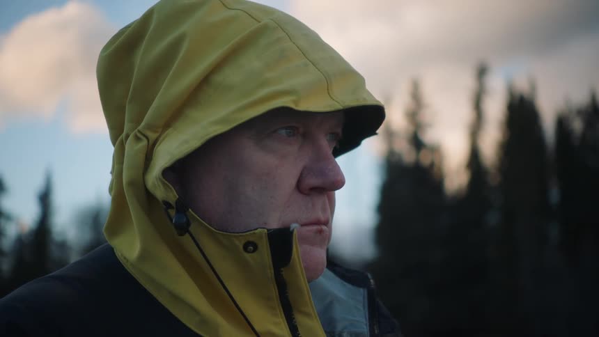 Man wearing jacket with emblem stands staring a short distance from conifer forest