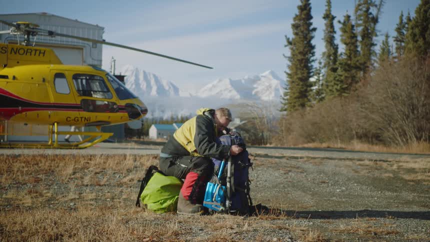 Man sits on one bag while looking through another bag in field near helicopter and hanger