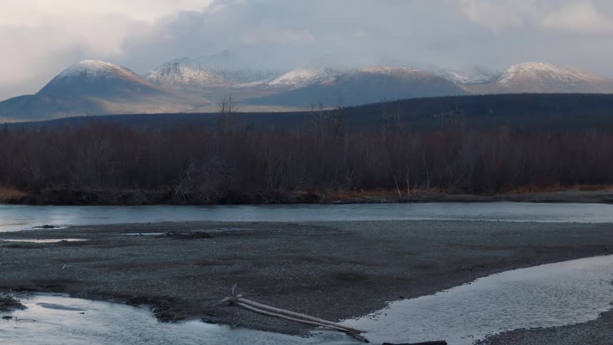 A flowing river in front of a bare forest in front of large mountains in the distance with snow covered peaks and fog rolling over them
