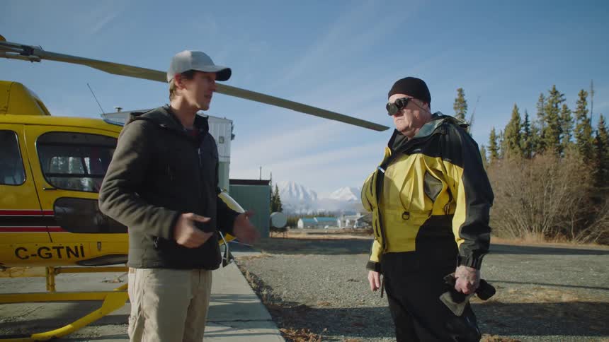 A man talks to a pilot in front of his yellow helicopter in a landing area near trees and mountains