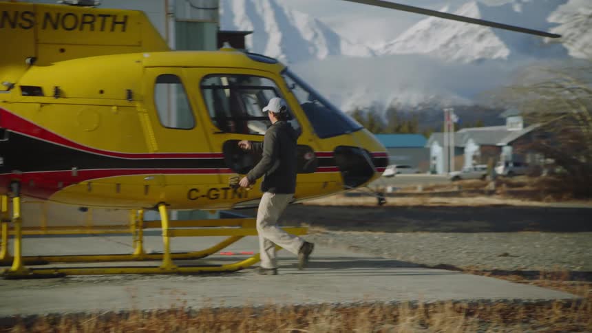 A man walks back to a yellow helicopter against a snow covered mountain