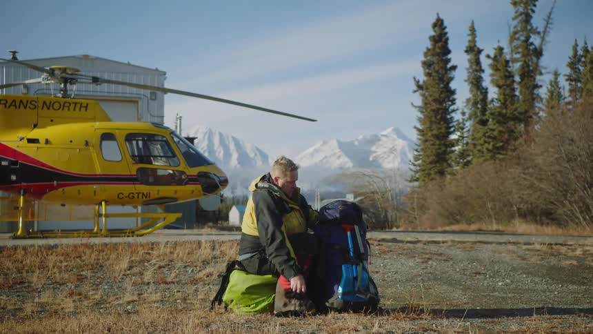 A man sits on the ground close to a helicopter and packs a backpack