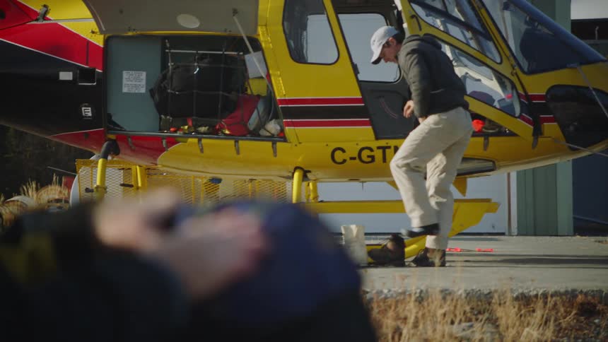 A man sits by a helicopter and packs a bag as another man gets boots out of the helicopter