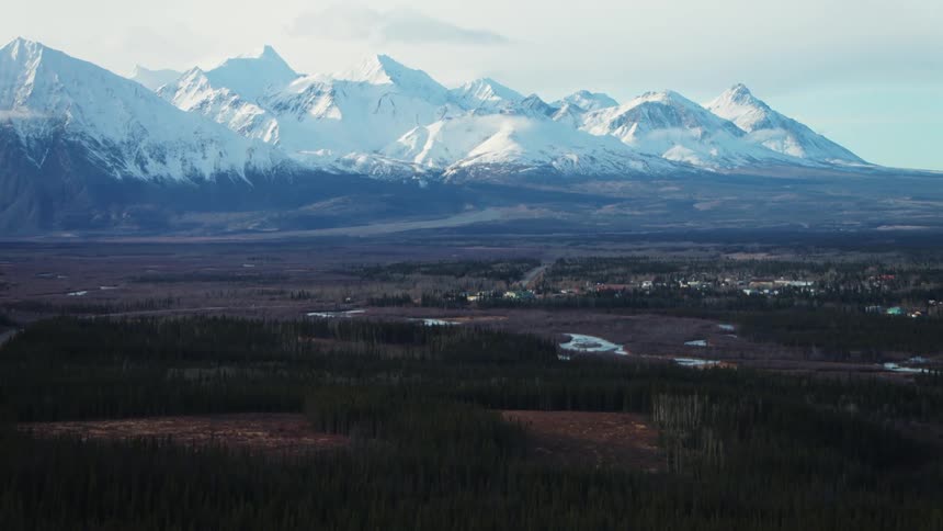 Orbiting aerial view of a small town in the plains next to the snow covered peaks of a mountain range at evening