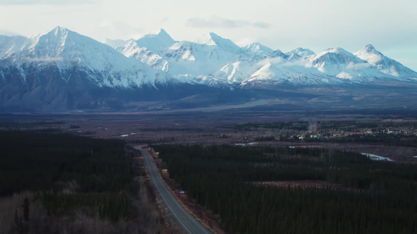 Road curves around woods as ice patches sit on fields near town with mountains in the background