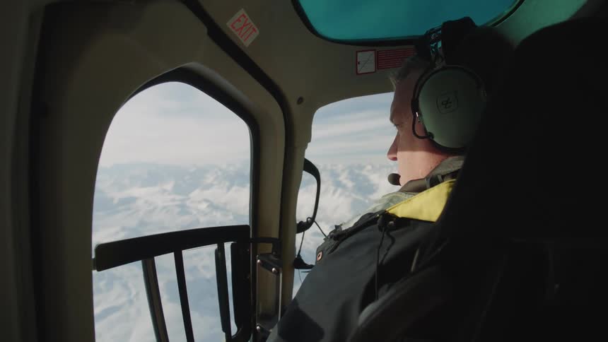 A man wearing a headset sits in a helicopter and looks out the window
