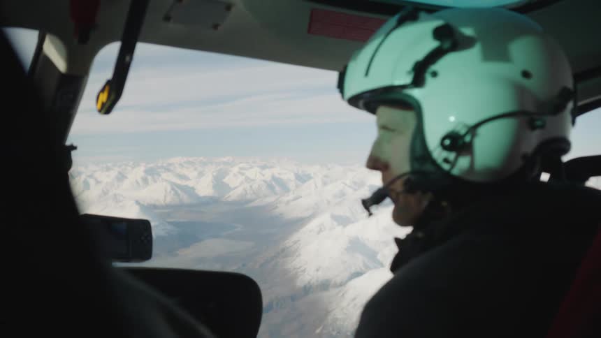 A helicopter pilot wearing a white helmet flying a helicopter over snowy mountains
