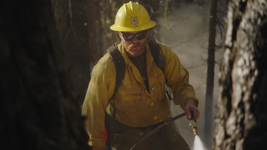 A man wearing a hardhat and sun glasses is standing near trees in the woods holding a water hose as smoke rises in the air