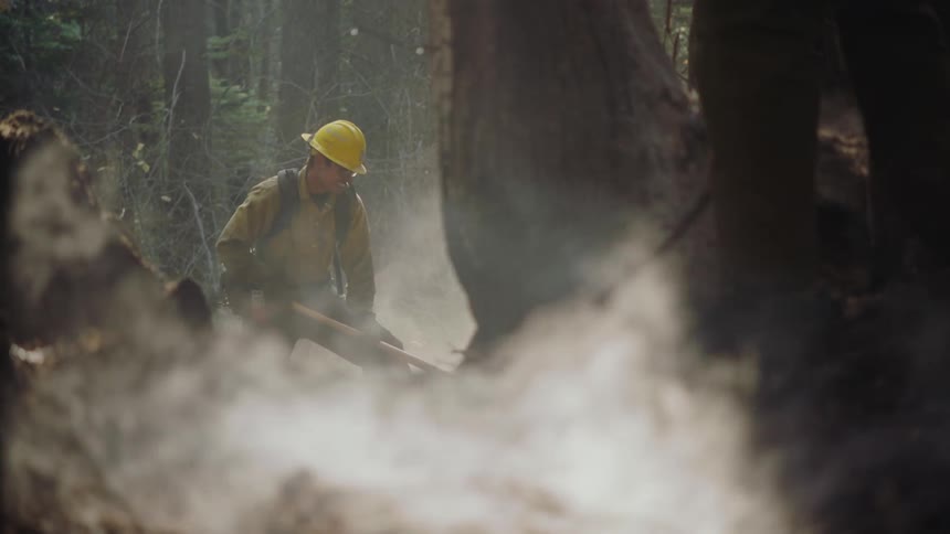 Firefighter wearing yellow protective gear surrounded by white smoke while shoveling in the forest
