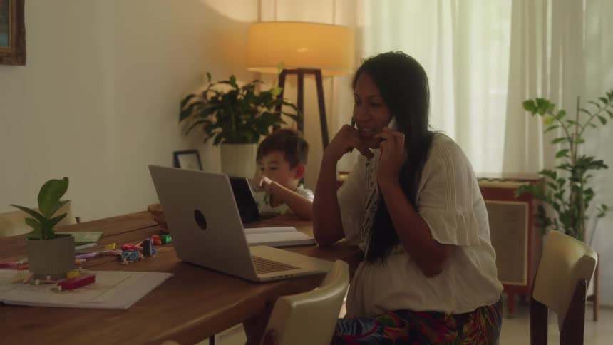 A woman is sitting at a table with a laptop computer writing as she talks on the phone and her little boy is looking at a tablet computer