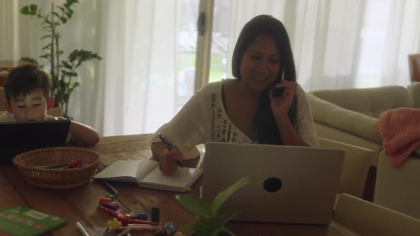 Boy views tablet while mother sits next to him at table and consults computer during a phone call