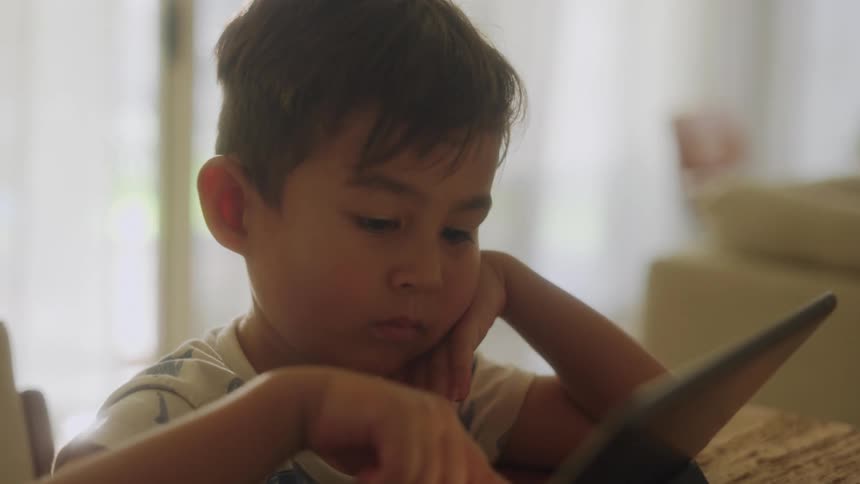 A boy is sitting in a tall chair at an island counter resting his cheek on the back of one hand and touching a tablet that is standing up on the counter in front of him with the other as he looks at the tablet with a board expression on his face
