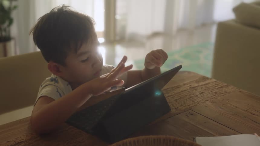A little boy is sitting at a table using both hands as he plays on a tablet computer