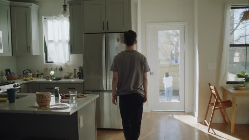Man arriving in kitchen walks to door to watch woman standing outdoors without coat in cold