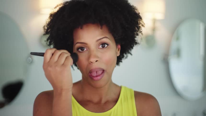 A woman uses a makeup brush to apply makeup to her face while looking into the camera as if it is a mirror