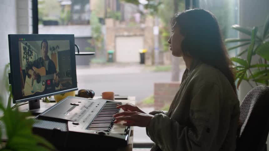 Woman plays keyboard in office while interacting with guitarist playing on monitor during a video call