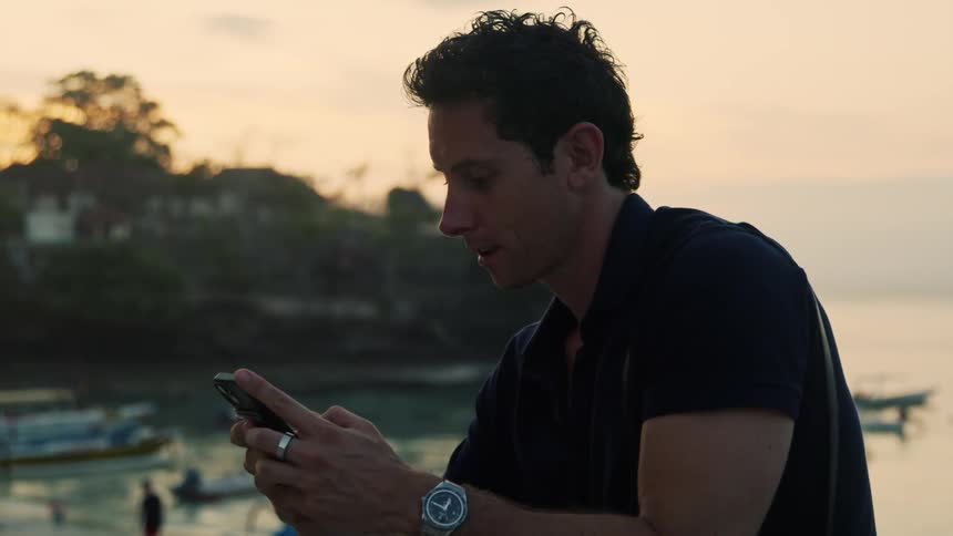 A man in a black tshirt looks at his phone while sitting near a dock