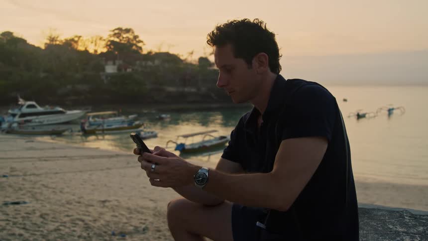 A man is sitting on the beach as he uses his cell phone and there are small boats in the water along the shore