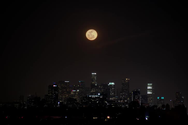 A time lapse view of a full moon rising into the night sky above a city skyline
