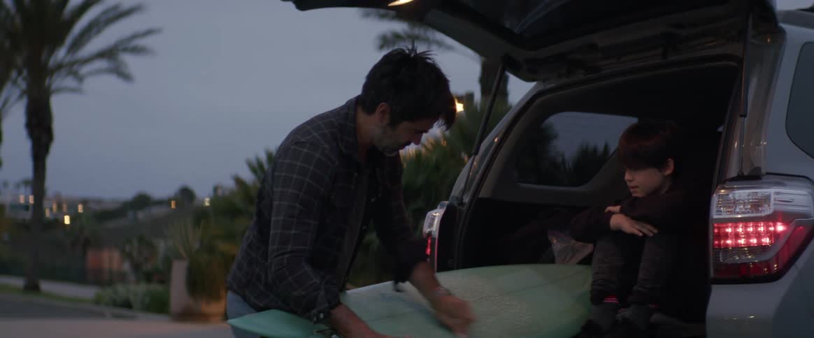Man waxes a surfboard on the trunk of a car next to his son at evening