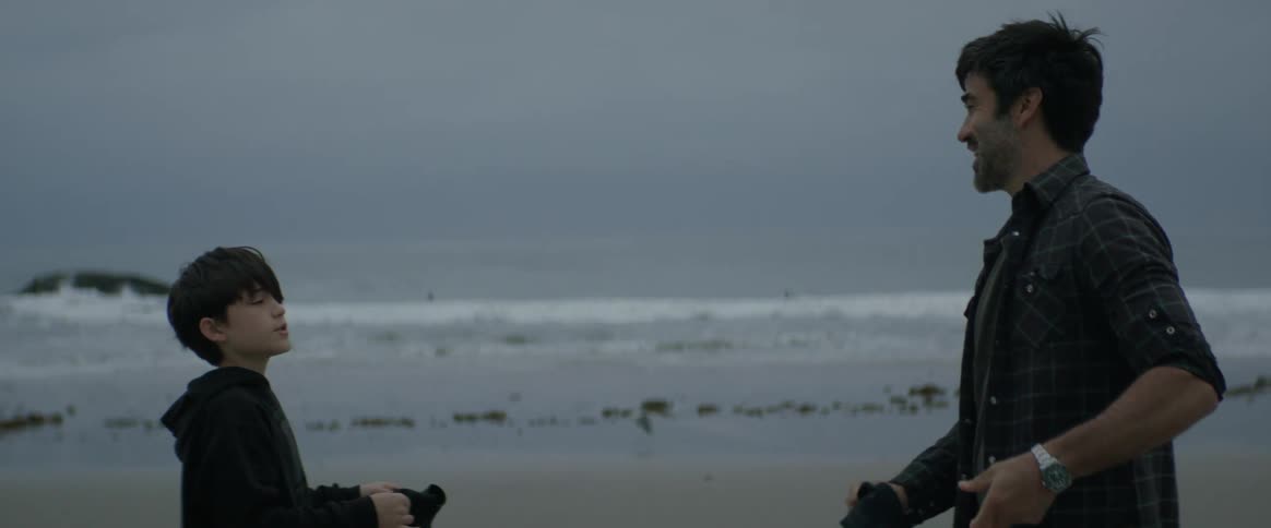 Father and son folding clothes while standing talking on the beach with the ocean behind them