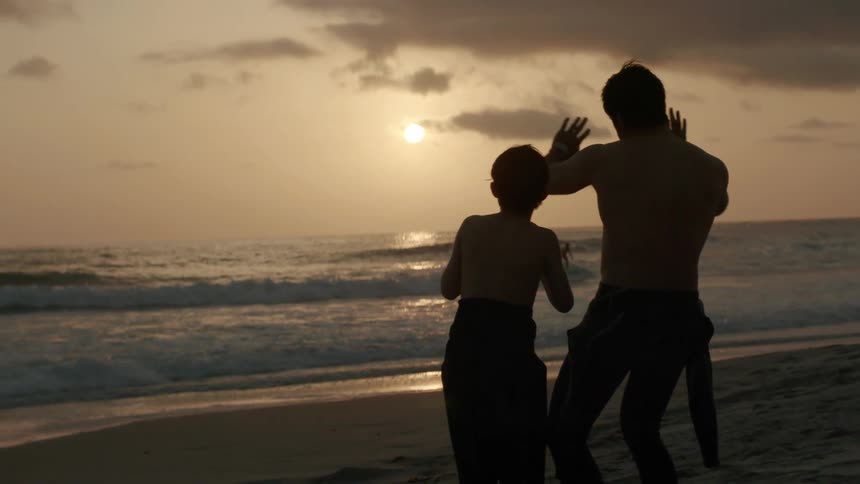 A man is on the beach with his young son as waves roll into shore and the sun sets in the distance