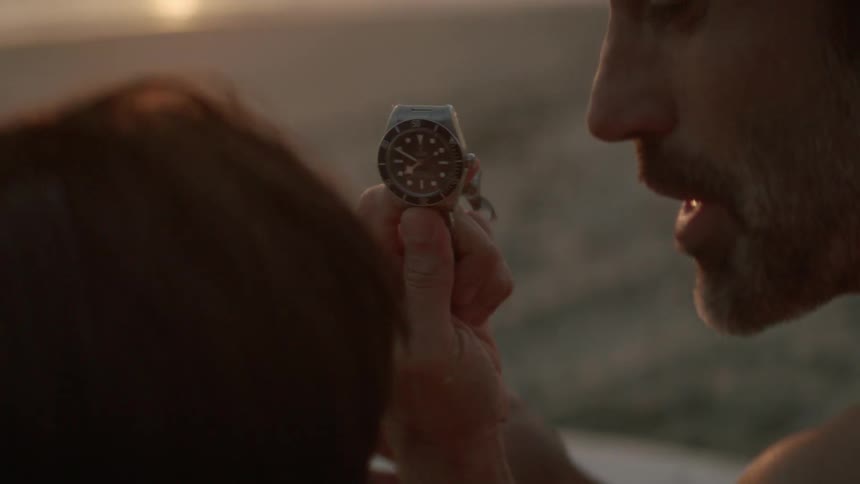 Topless father with beard giving silver wristwatch to his son on the beach at sunset