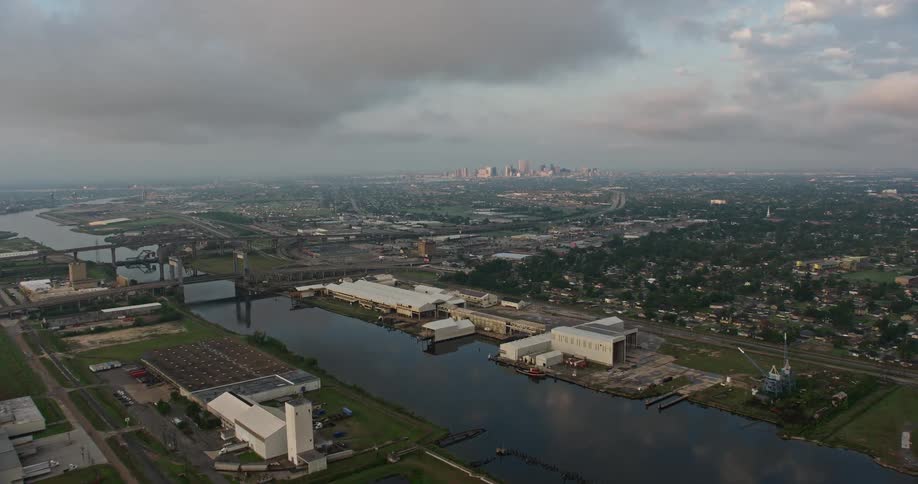 A stunning 4K aerial footage at sunrise captures the Industrial Canal in Upper Ninth Ward New Orleans as bridges stretch across the river near the warehouses amidst a beautiful skyline and cloudy skies