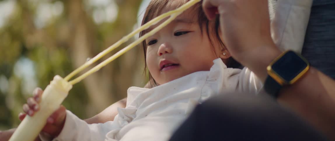 A young asian girl sits outdoors on her father's lap while they play with a soap bubble toy