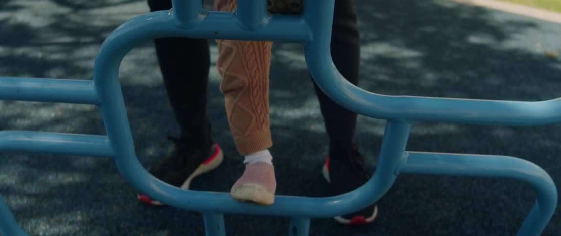 A father holding his toddler daughter as she climbs blue bars on a playground