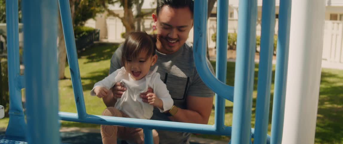 A father lifts his daughter onto a playground platform smiling and laughing