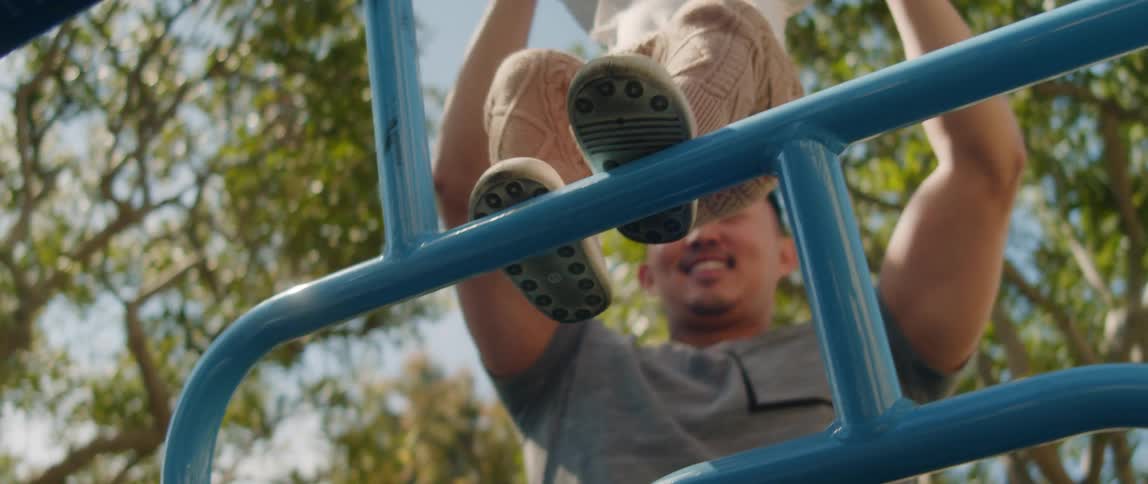 A father holds his toddler daughters hands and helps her climb blue bars on a playground