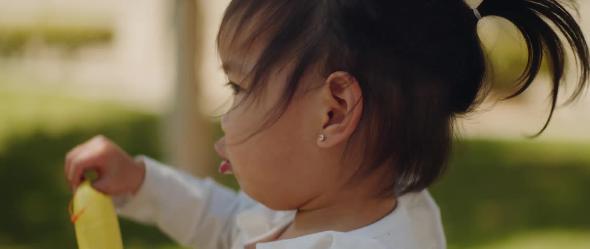 Little baby toddler girl sitting outside in the grass with the wind blowing in her hair smiling and playing with a toy looking back at daddy