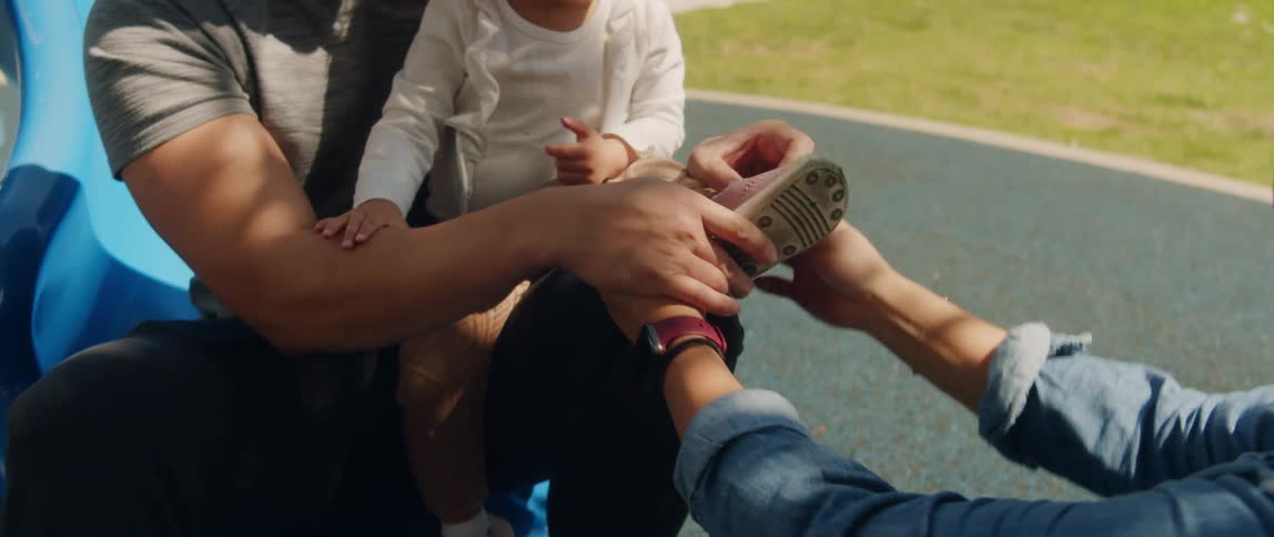 Father sits on playground slide holding daughter as mother puts shoe onto her foot