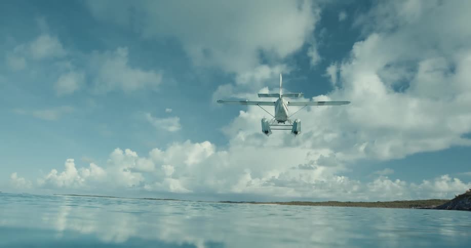 Reveal from under water of seaplane taking off over blue water into blue sky with puffy clouds