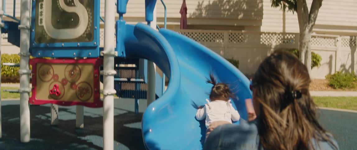 Mother claps while her daughter uses a slide on a swing set in the backyard at morning