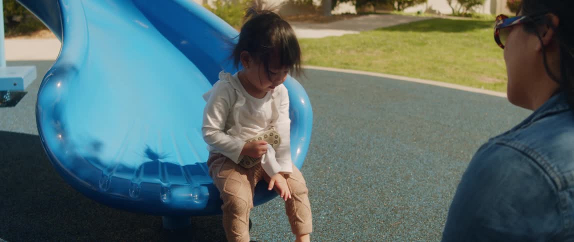 A child sits on a blue slide on a playground and a woman pushes hair out of their face