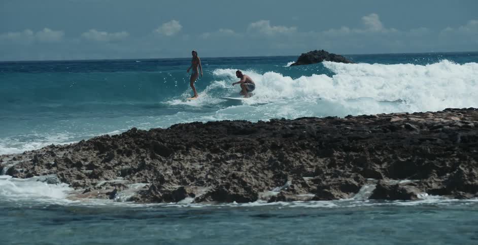 A man and a woman surfing side by side in the blue ocean behind black volcanic rock