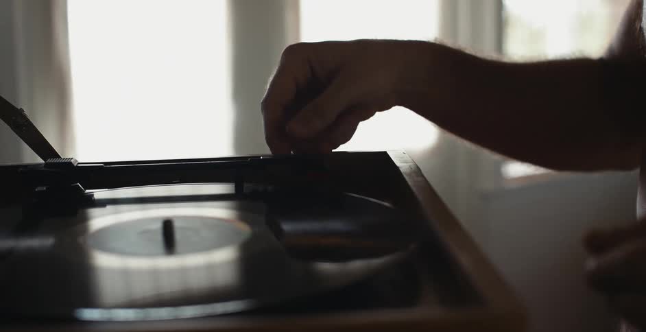 A man walking to a record player and putting a disc in it and putting the needle on the record to play it