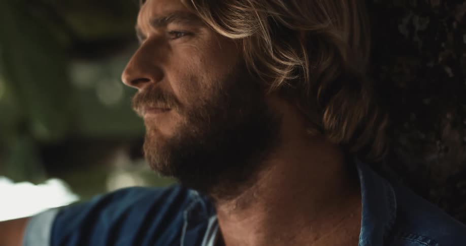 A close up of a man sitting playing a guitar looking straight ahead