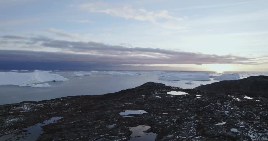 An aerial of the arctic ocean coastline meeting rocky mountains with large icebergs in the ocean