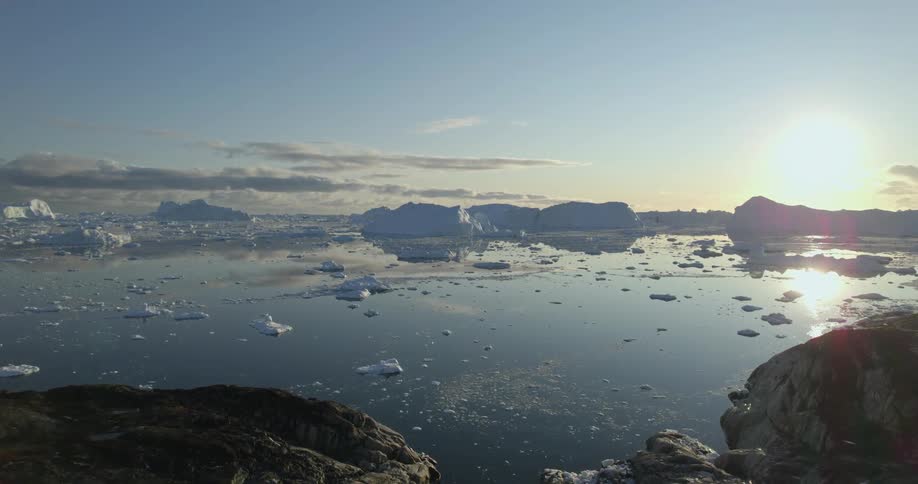 An aerial of the arctic ocean with rock coastlines and large icebergs as the sun reflects off the water