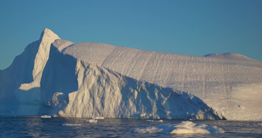 The sun shines brightly as pieces of ice float among icebergs in the water under a clear blue sky