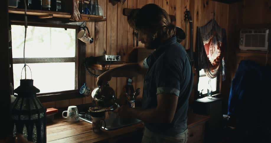 Man pours hot water through ground coffee into mug while standing in wooden paneled kitchen