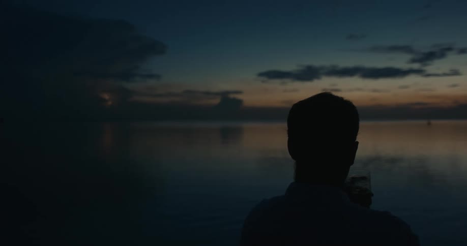 A mans silhouette from the back as he looks out over the ocean as a lightning storm flashes over