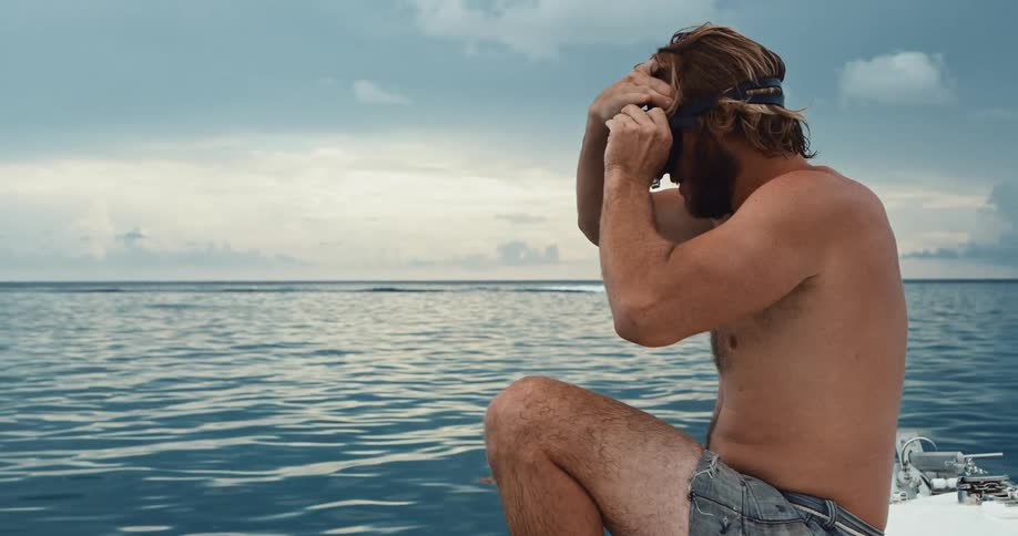 A man sitting on the edge of a boat putting goggles on as a boat drives by behind him