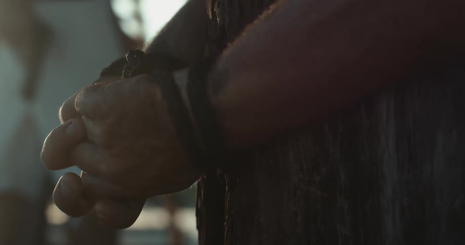 Person holding pistol walks past man tied to wooden tree trunk