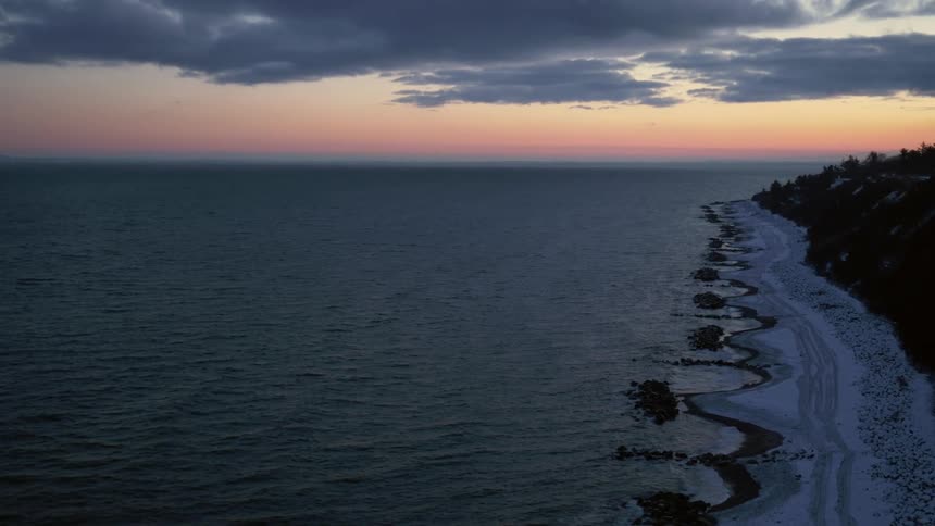An aerial landscape view of ocean waves crashing into a snowcovered beach near a forest at dusk