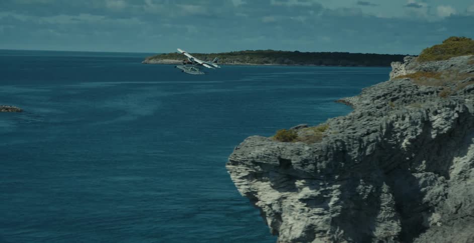 Aerial view of rocky coast revealing seaplane taking off from island over blue water toward another island